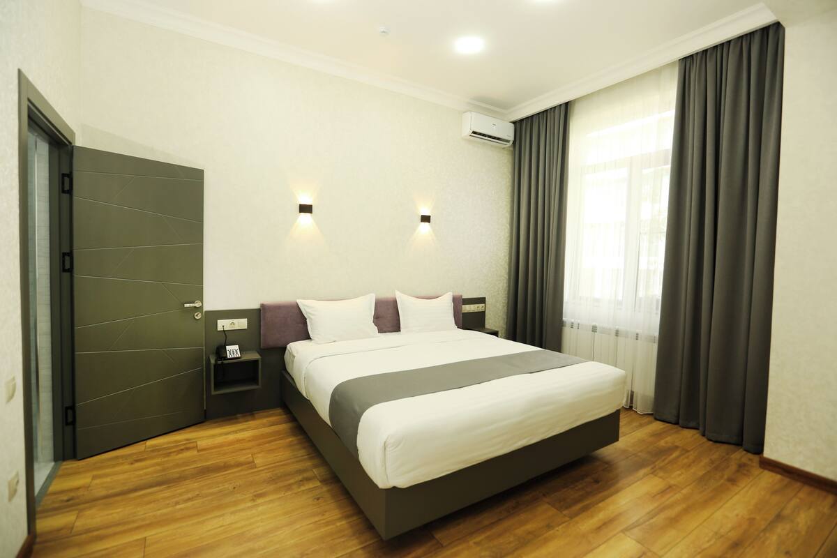 A modern hotel room with a large bed, a bedside table, an air conditioning unit, and a window with curtains.
