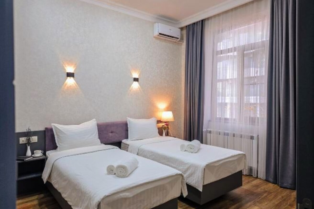 A hotel room with two beds, a nightstand, a lamp, and a window with curtains.