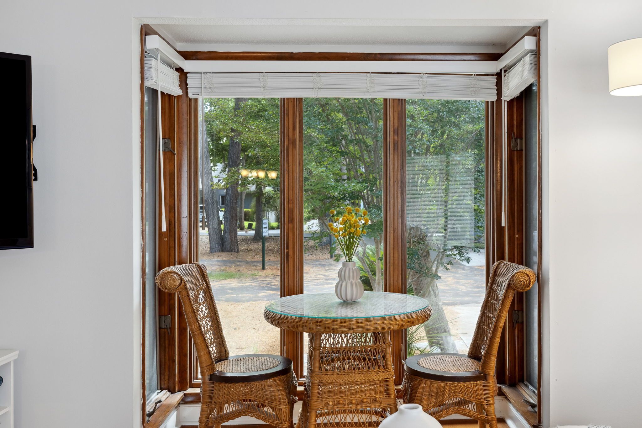 A room with a glass table and wicker chairs, a vase with yellow flowers, and a view of trees and a pathway outside.
