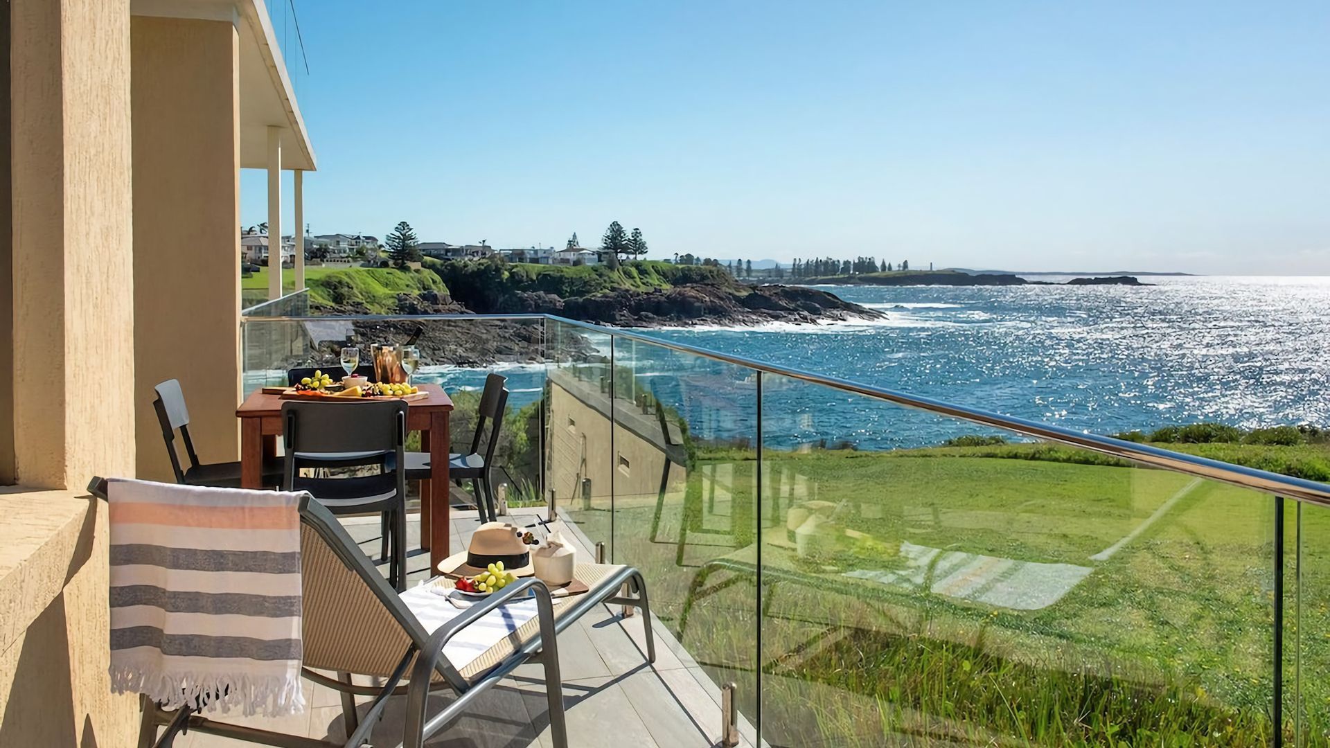A balcony with a table set for dining, overlooking a coastal view with the ocean and greenery.