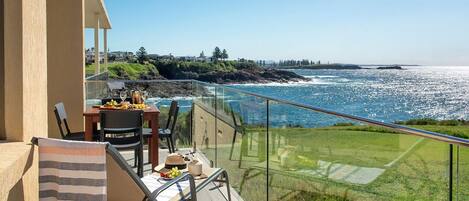 Un balcón con una mesa lista para comer, con vista al mar y vegetación costera.