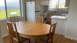 A dining area with a wooden table and chairs, a white refrigerator, and a view of the ocean. - Headland Hideaway ~ Spectacular views for whale watching! (Kiama)