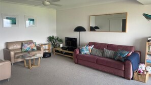 A living room with a beige sofa, a red sofa, a glass coffee table, a TV, and a bookshelf. - Headland Hideaway ~ Spectacular views for whale watching! (Kiama)