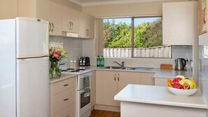 A modern kitchen with white cabinets, a stove, oven, and a sink. There is a window with a view of greenery outside. - Headland Hideaway ~ Spectacular views for whale watching! (Kiama)