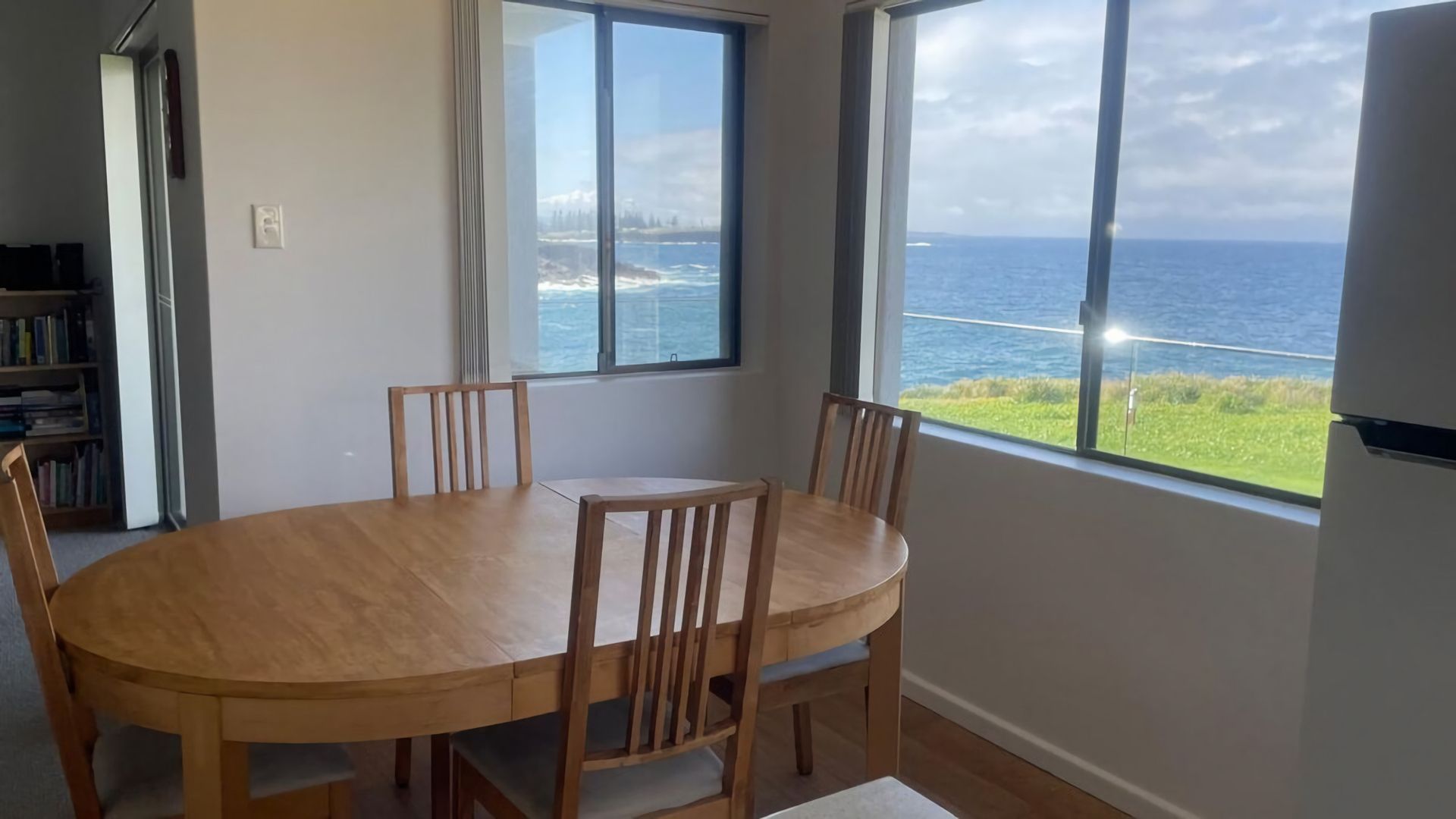 A dining area with a round table and chairs, large windows with a view of the sea, and a bookshelf in the background.