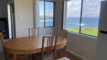 A dining area with a round table and chairs, large windows with a view of the sea, and a bookshelf in the background.