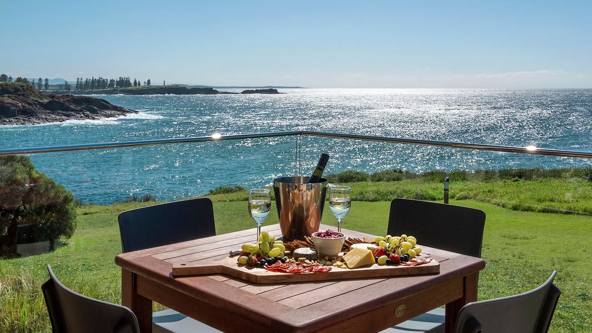 An outdoor dining area with a view of the ocean, a wooden table with a variety of cheeses and fruits, and a bottle of champagne.