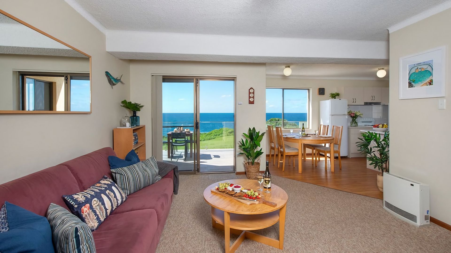 A living room with a red sofa, a dining table with chairs, a kitchen area, and a sliding glass door leading to a balcony with a view of the sea.
