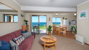 A living room with a red sofa, a dining table with chairs, a kitchen area, and a sliding glass door leading to a balcony with a view of the sea.