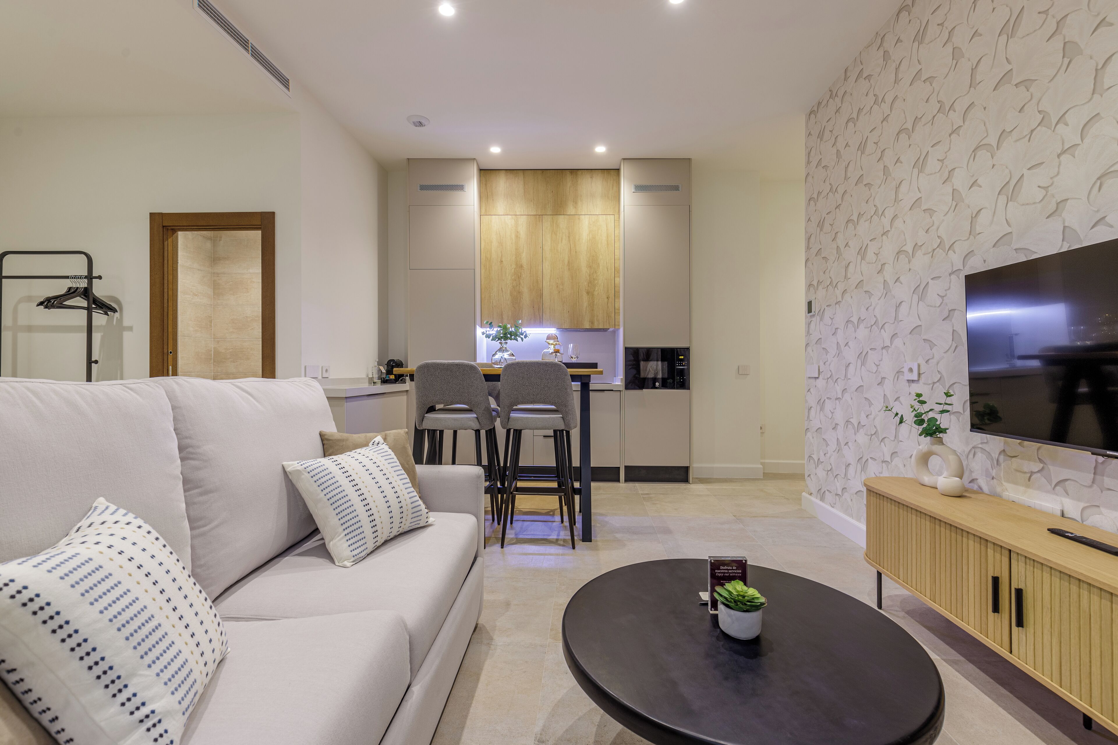 Una sala moderna con sofá, mesa de centro y televisor. Se observa un área de cocina con gabinetes de madera y una mesa de comedor.