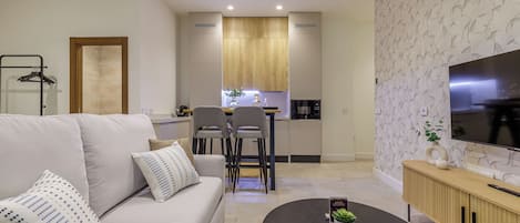 Una sala moderna con sofá, mesa de centro y televisor. Se observa un área de cocina con gabinetes de madera y una mesa de comedor.
