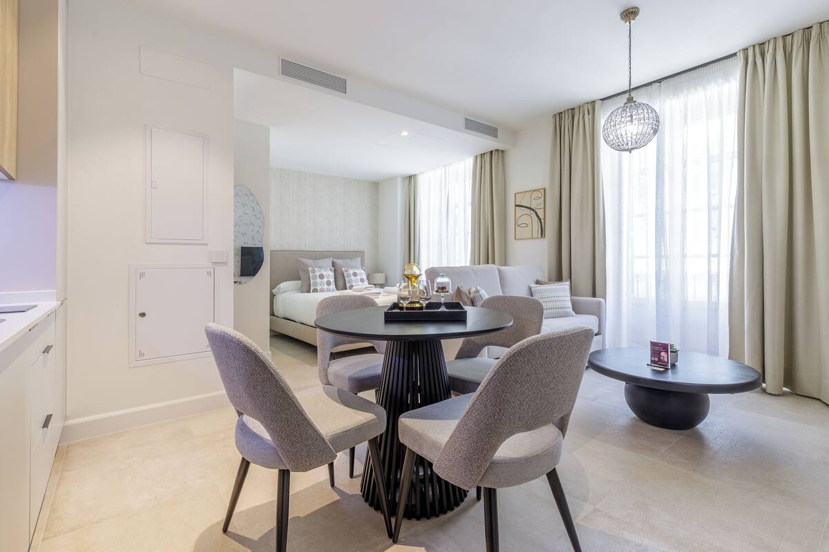 Une chambre d’hôtel moderne avec une table à manger ronde, quatre chaises grises, un canapé et une grande fenêtre avec des rideaux.