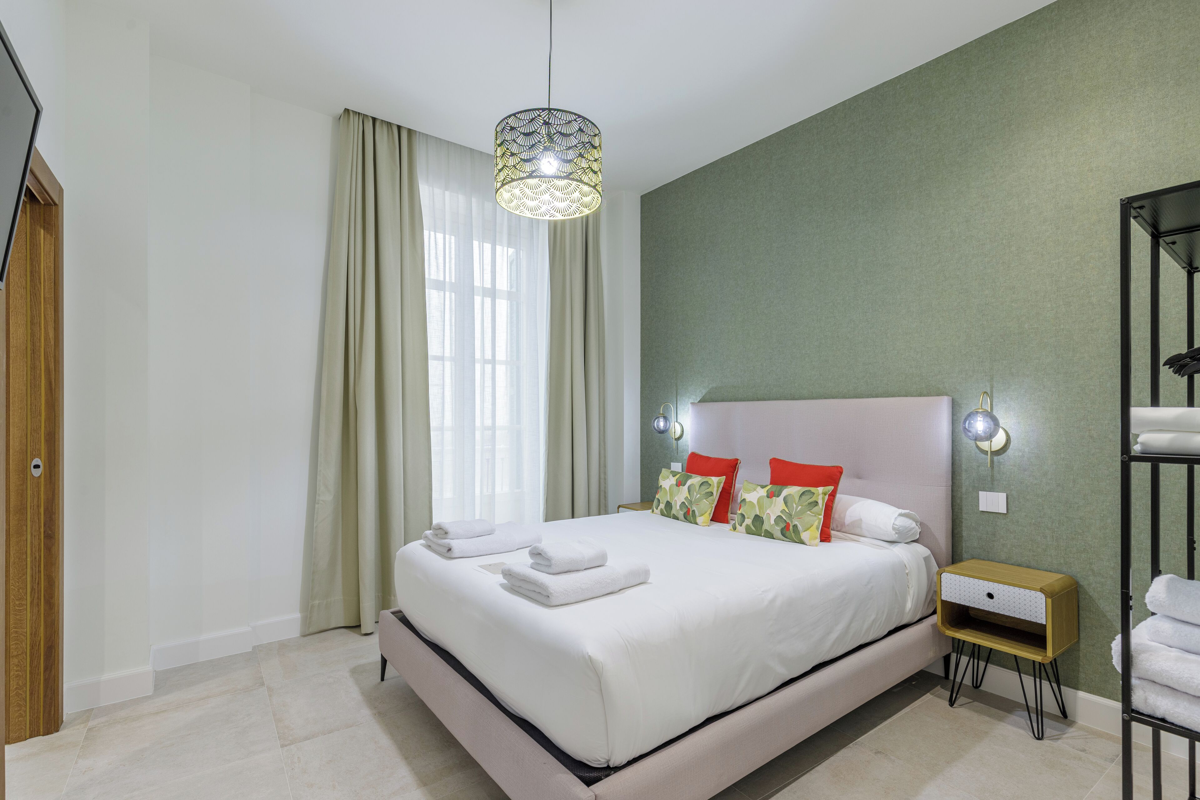 A neatly made bed with white linens and towels, a bedside table with a lamp, and a view of the window with curtains.