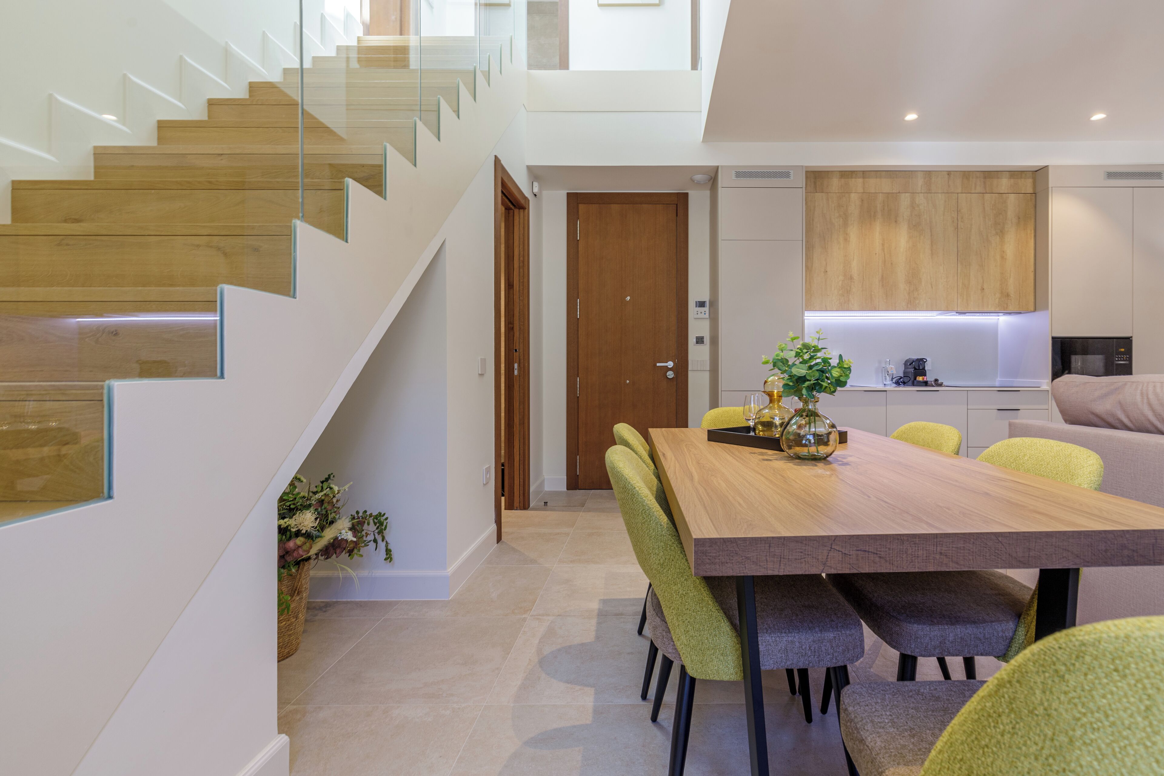 A modern kitchen with wooden cabinets, a dining area with a wooden table and chairs, and a staircase with glass railings.