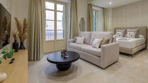 A modern living room with a sofa, a coffee table, and a bed. There are large windows with curtains, a mirror, and a plant on the side table. - Plaza del Siglo Suites (Málaga)