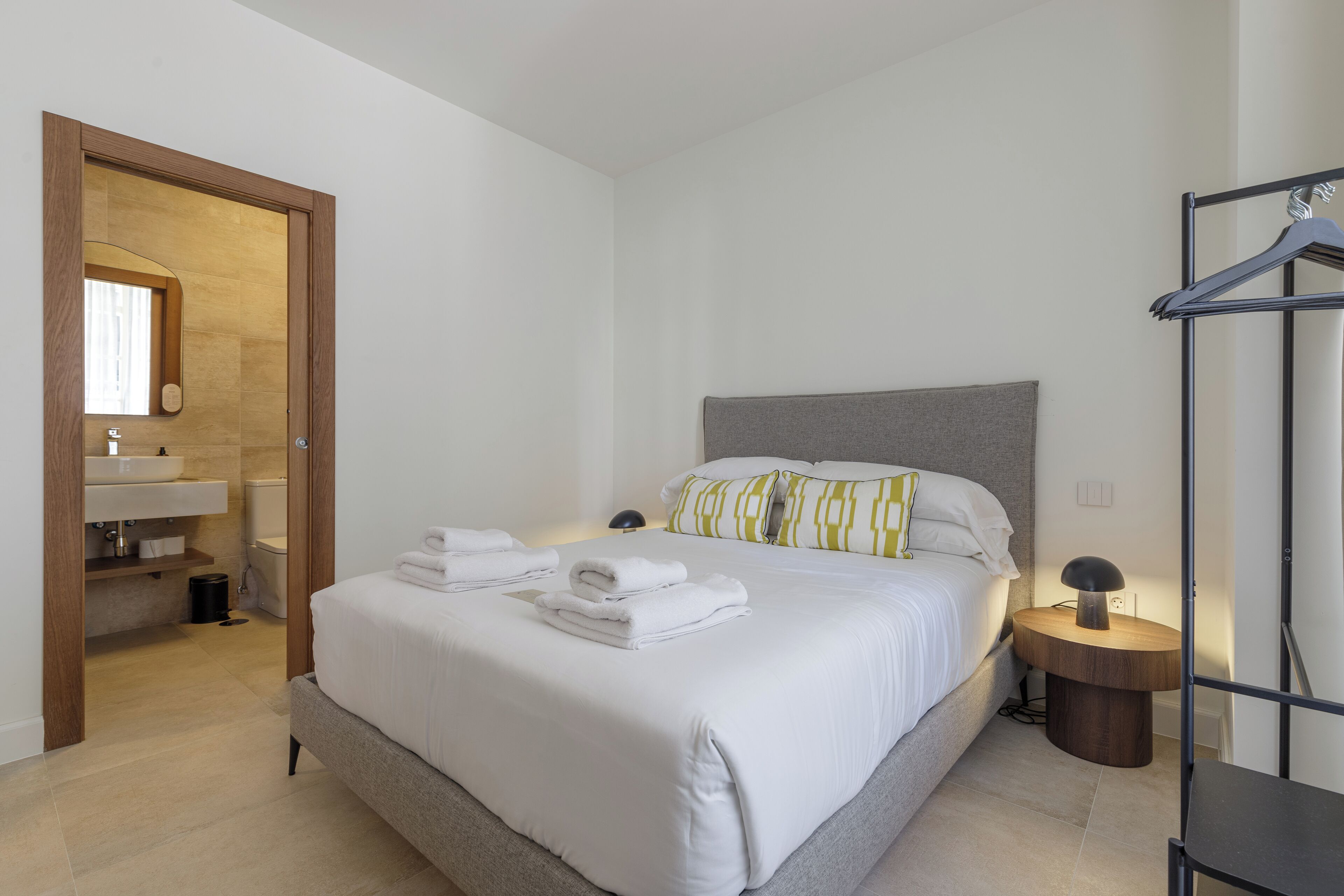 A neatly arranged bedroom with a bed, bedside tables, a hanging clothes rack, and a bathroom with a sink and mirror.