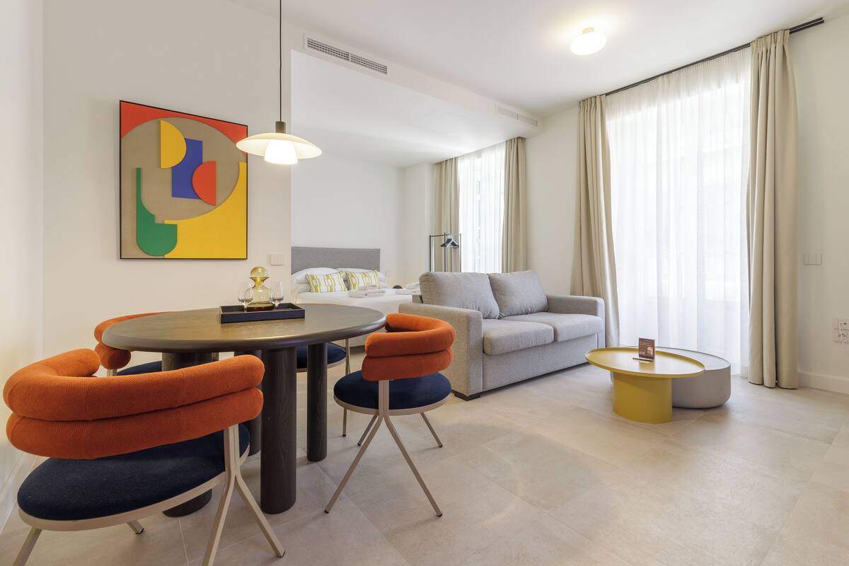 Un salon moderne avec une table de salle à manger ronde, des chaises orange et bleues, un canapé gris et une table basse jaune.