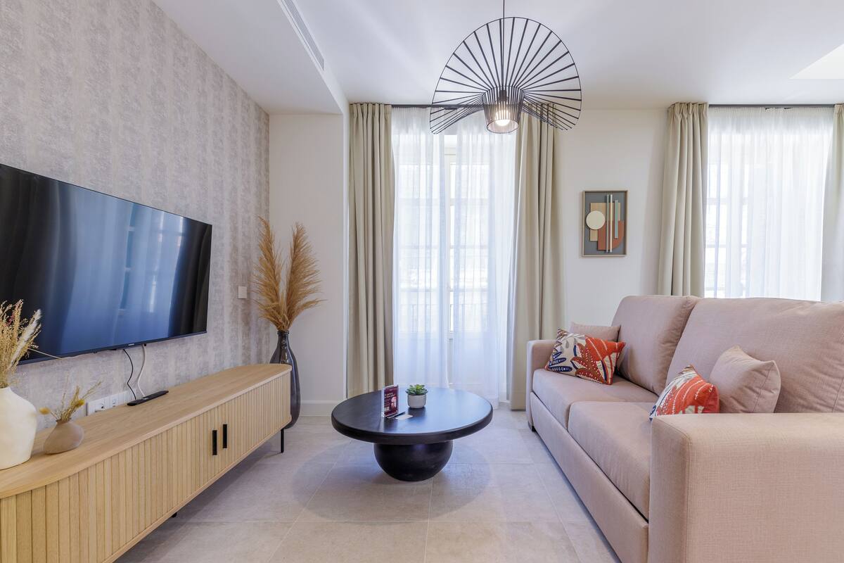 Un salon moderne avec un téléviseur à écran plat, une table basse ronde, un canapé beige et une grande fenêtre avec des rideaux.