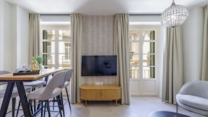 A modern living room with a flat-screen TV mounted on the wall, a wooden coffee table, a grey sofa, and a dining area with chairs and a table. - Plaza del Siglo Suites (Málaga)
