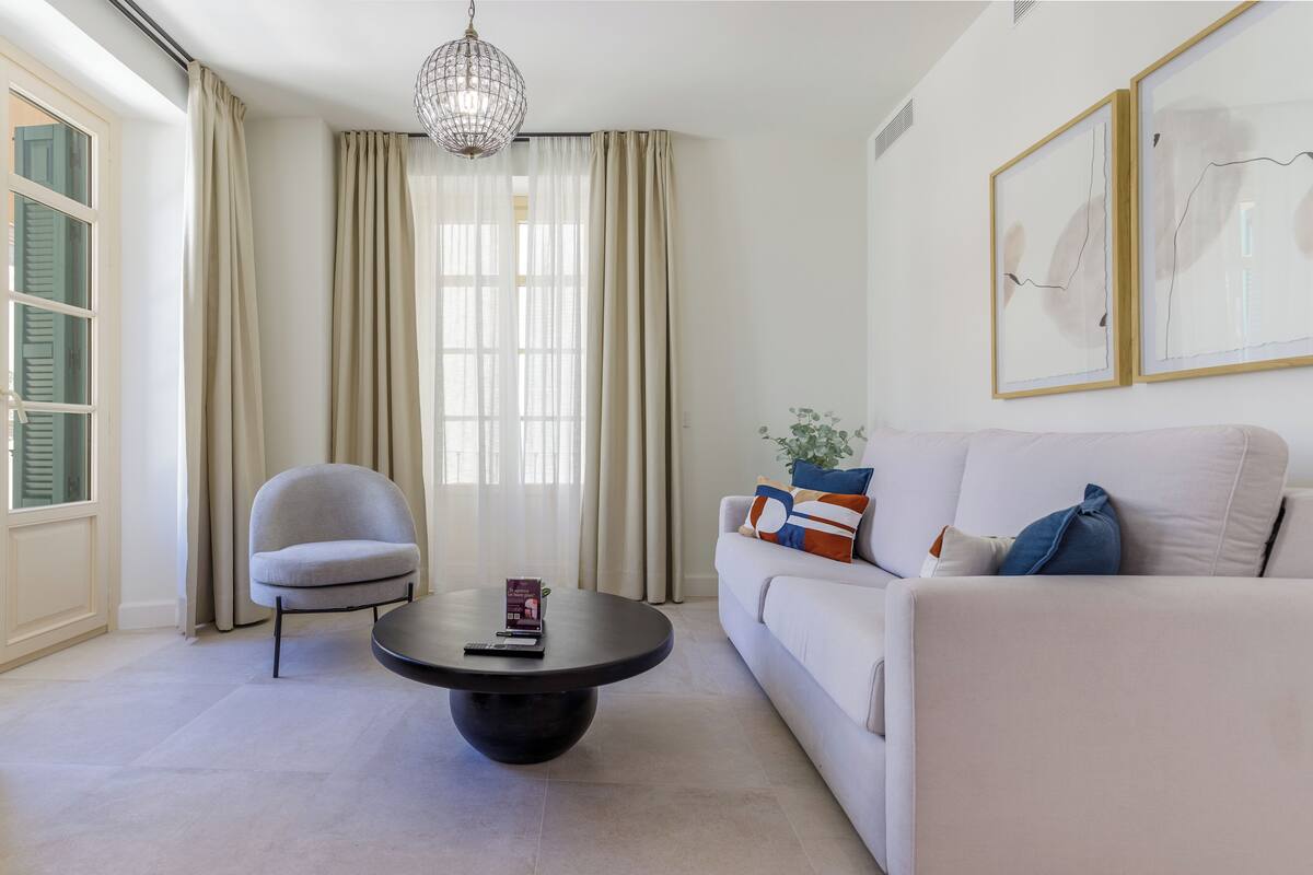 Un salon moderne avec un canapé blanc, une table basse ronde, un fauteuil gris et une grande fenêtre ornée de rideaux légers.