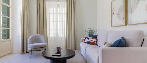 A modern living room with a white sofa, a round coffee table, a grey armchair, and a large window with sheer curtains.