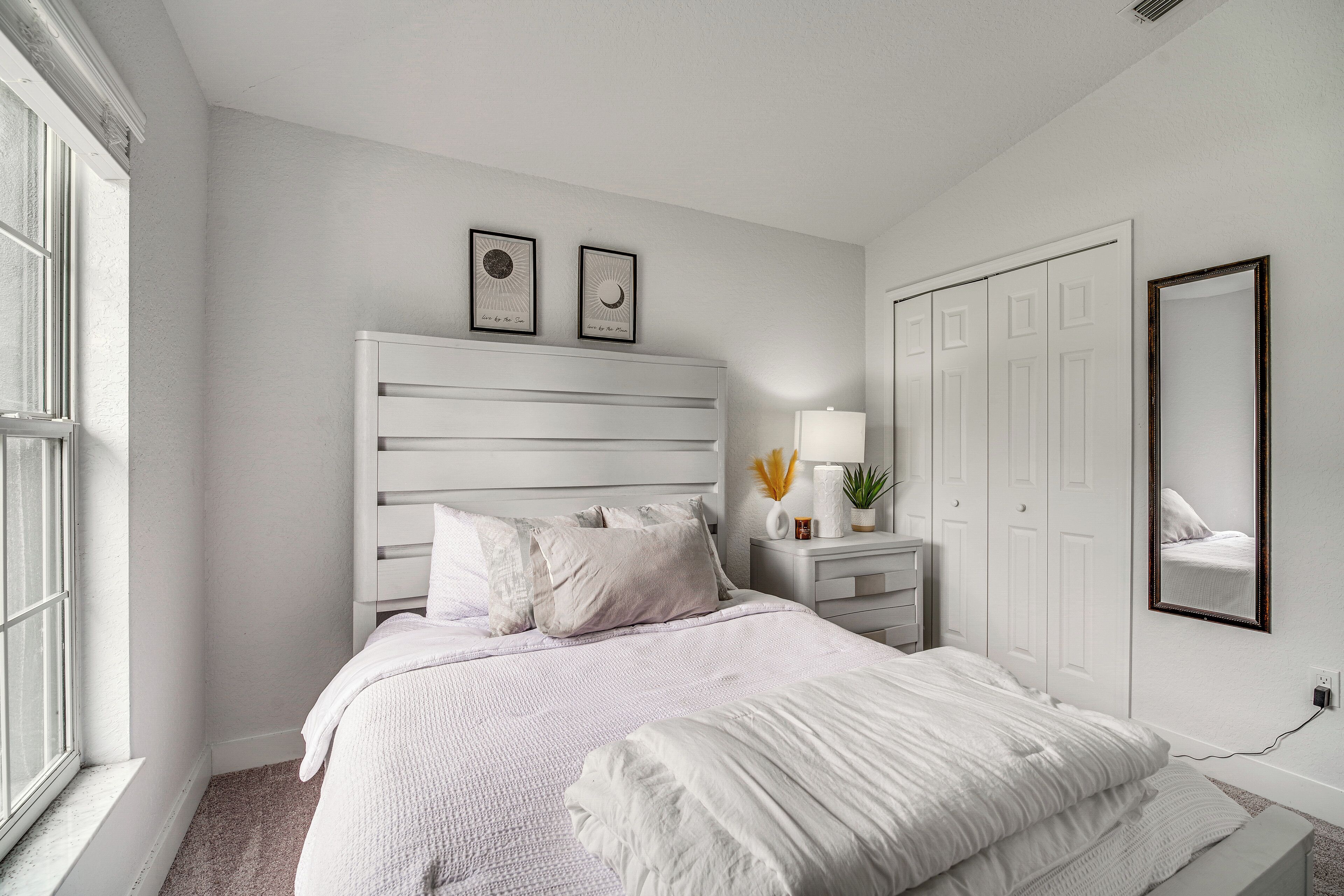 A bedroom with a white bed, a nightstand with a lamp, a dresser, and a large mirror on the wall.