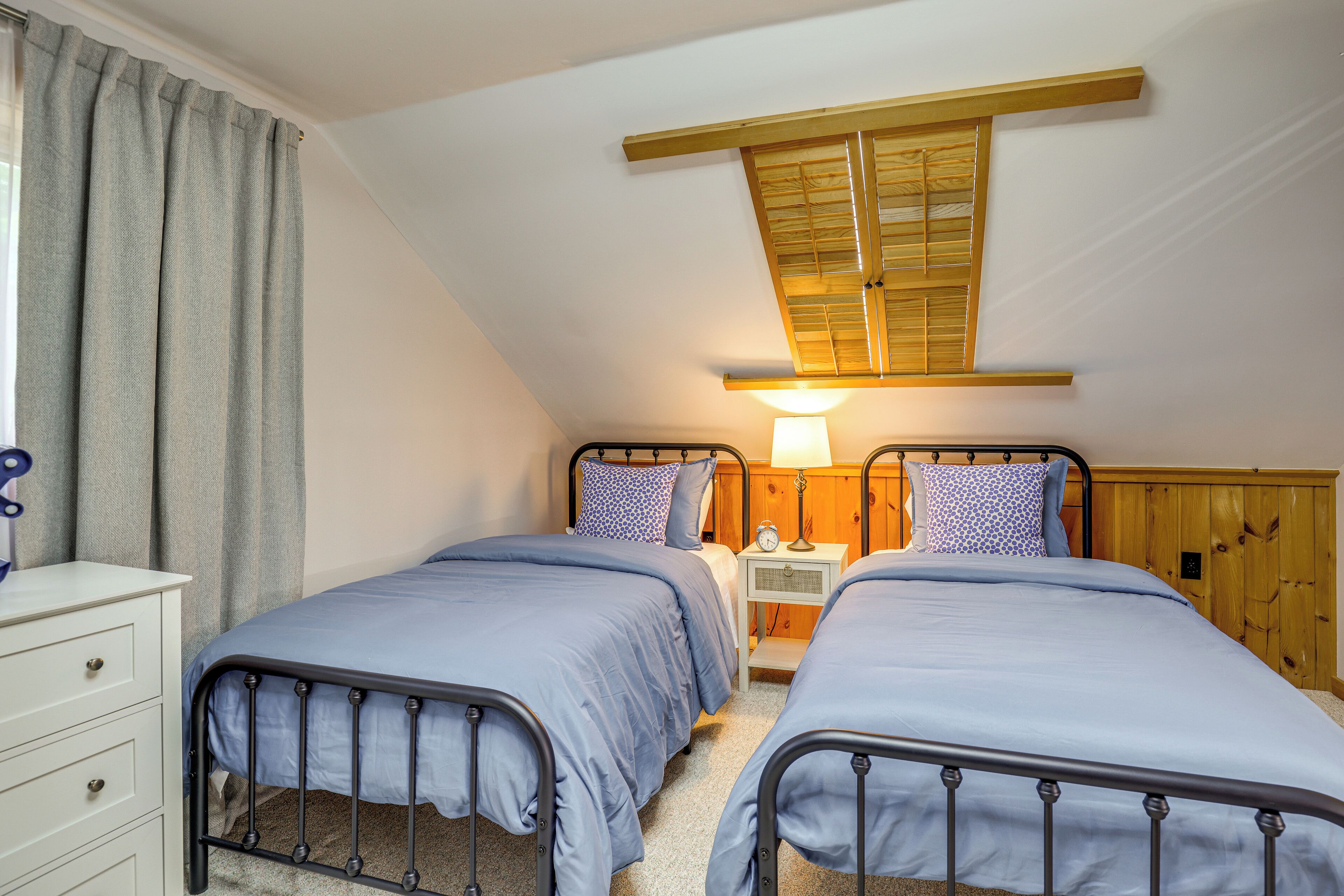 A bedroom with two beds, wooden headboard, a nightstand with a lamp, and a window with wooden shutters.