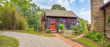 A rustic cabin with a red door, surrounded by greenery and a gravel path leading to it.
