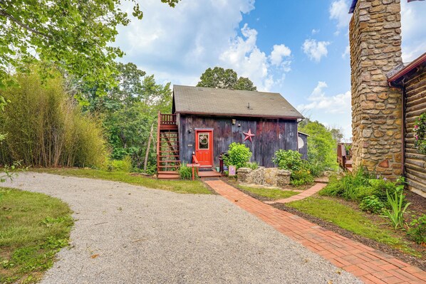 A rustic cabin with a red door, surrounded by greenery and a gravel path leading to it.
