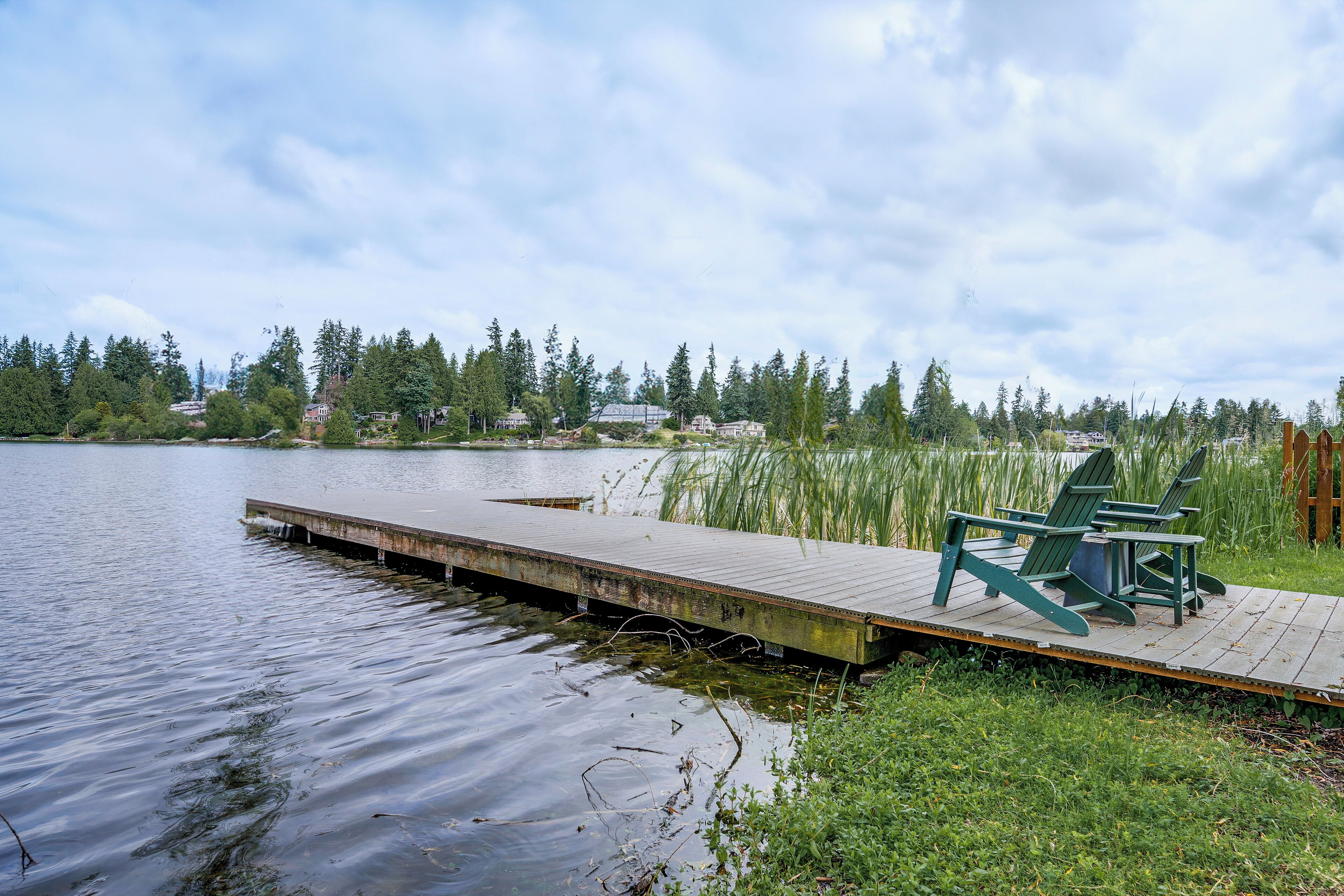 A wooden dock extending into a calm lake, with a grassy area and chairs on the right side.