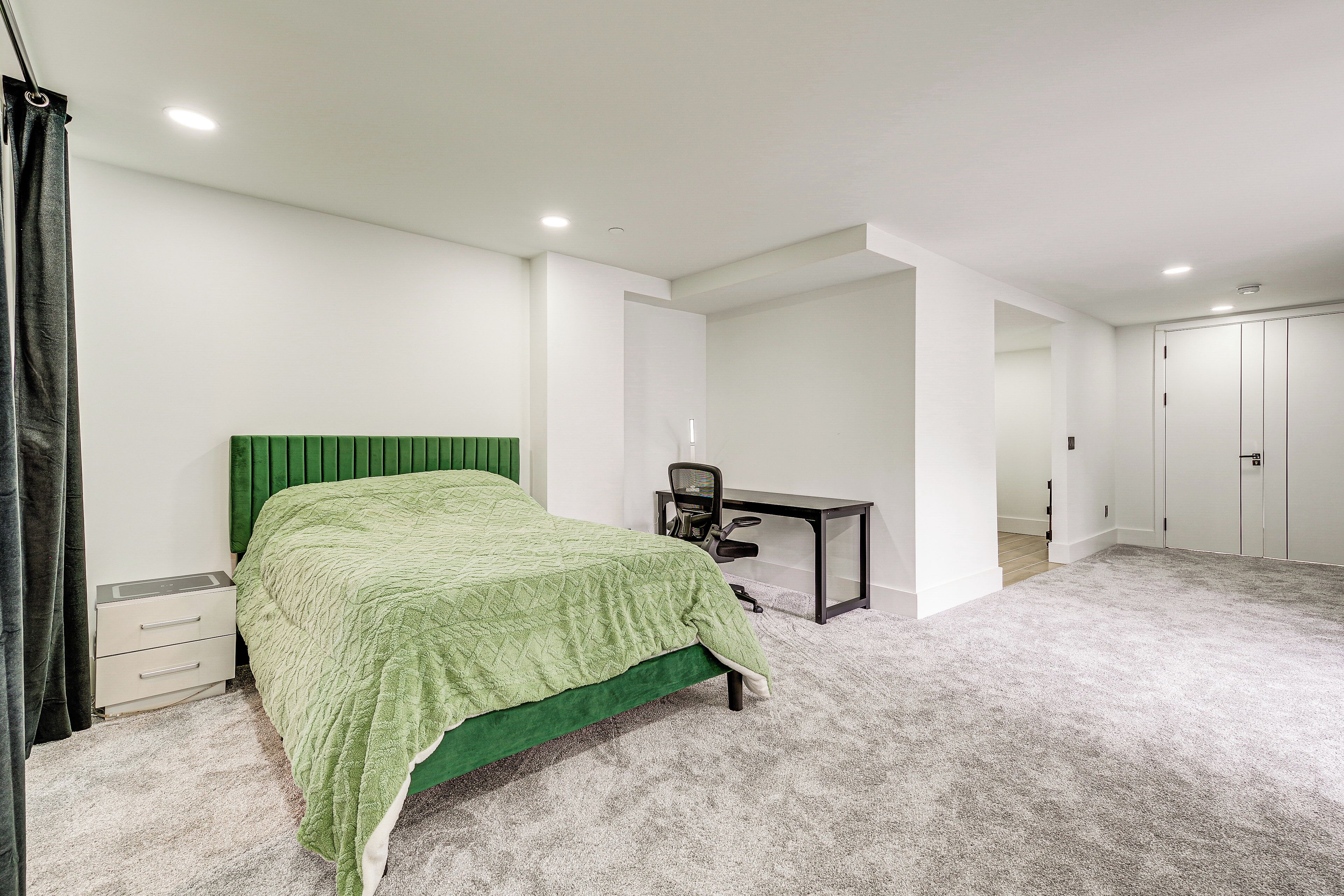 A bedroom with a green bed, a desk, and a chair. There are white walls and a carpeted floor.