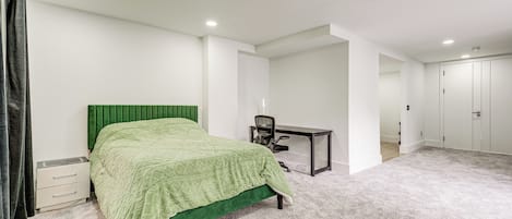 A bedroom with a green bed, a desk, and a chair. There are white walls and a carpeted floor.