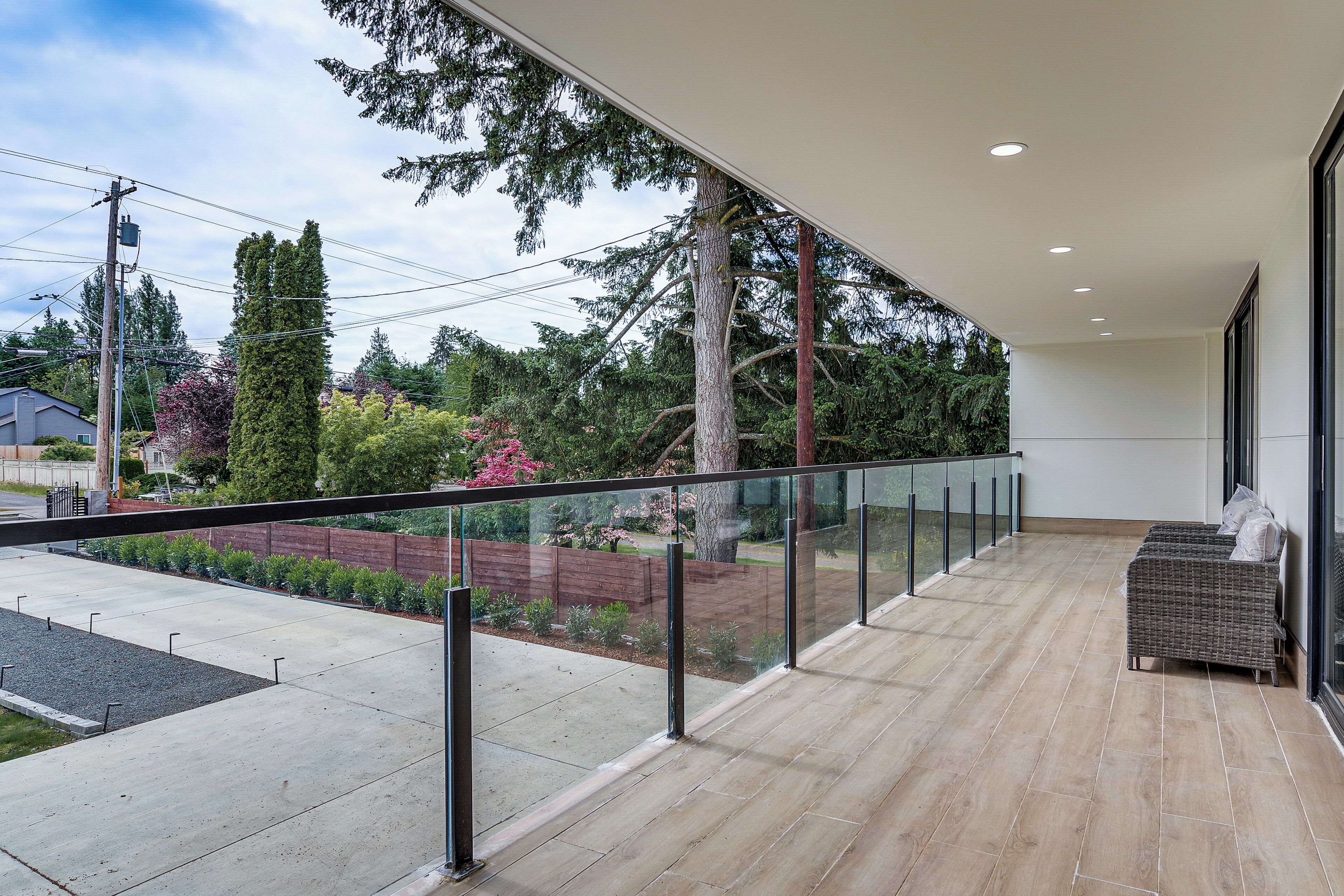 A modern balcony with a glass railing, a wicker sofa, and a view of a garden and neighboring houses.