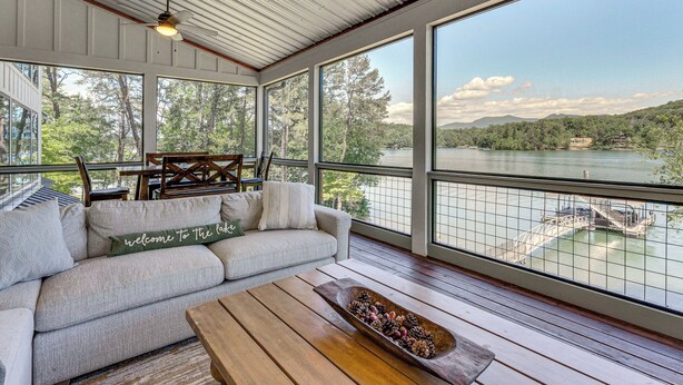 Ein geräumiges Zimmer mit großem Fenster mit Blick auf einen See, einer Holzdielen-Terrasse und einer gemütlichen Sitzecke mit Sofa und Couchtisch.