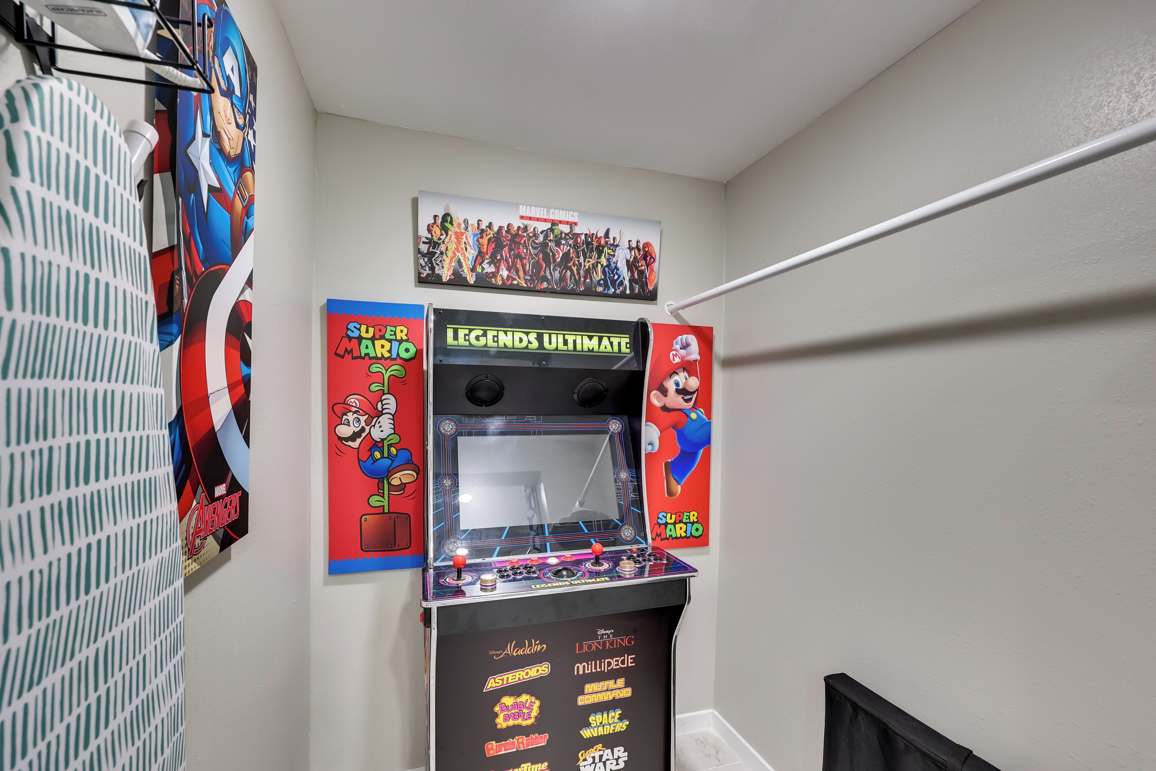 A room with a gaming arcade featuring 'LEGENDS ULTIMATE' and 'Super Mario' games, and posters of Marvel characters on the wall.