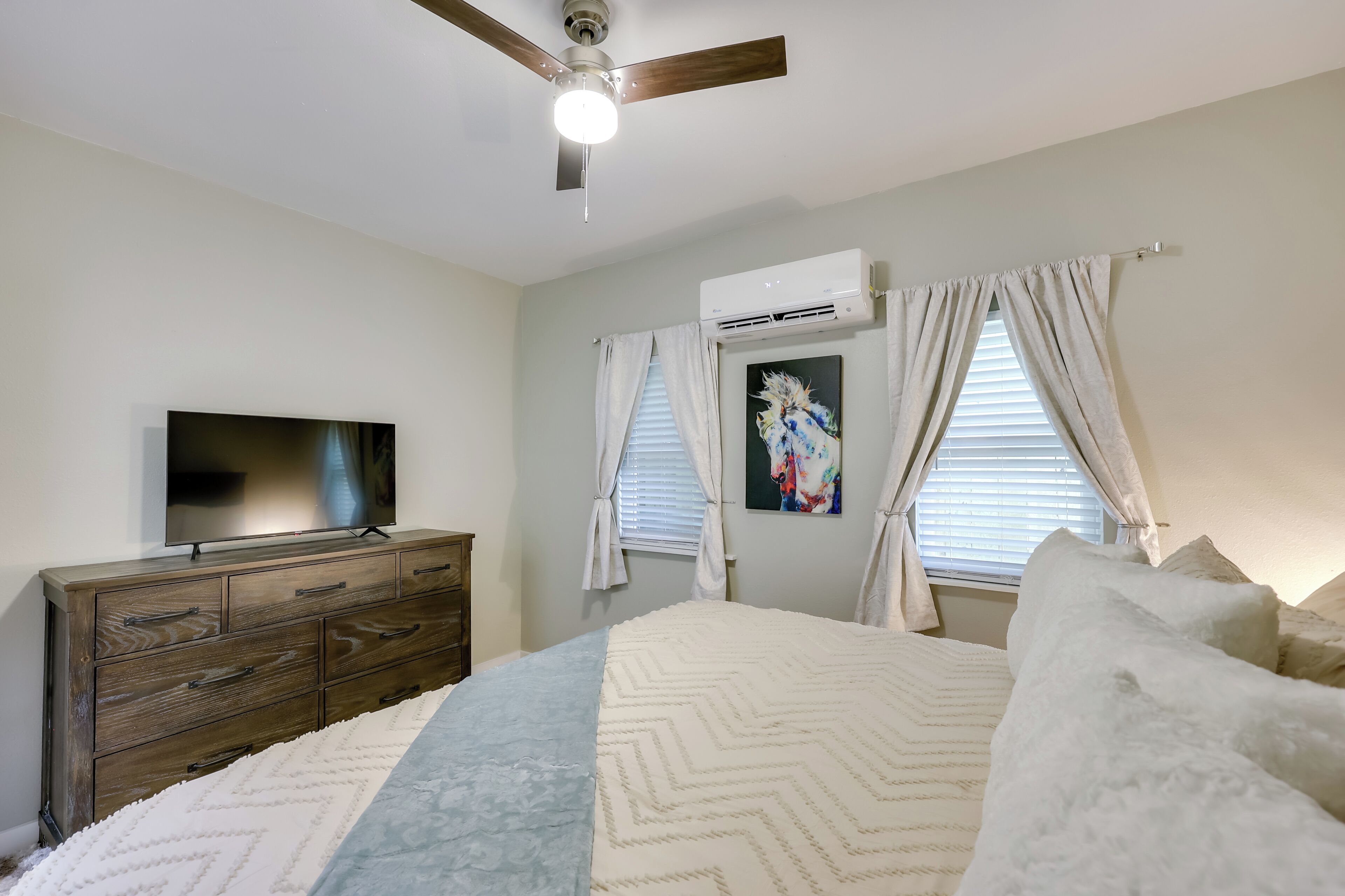A bedroom with a bed, a dresser, a ceiling fan, an air conditioner, and a TV.