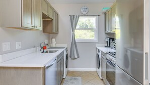 A compact kitchen with a built-in oven, a dishwasher, and a refrigerator. There is a window with a curtain, and a clock on the wall.