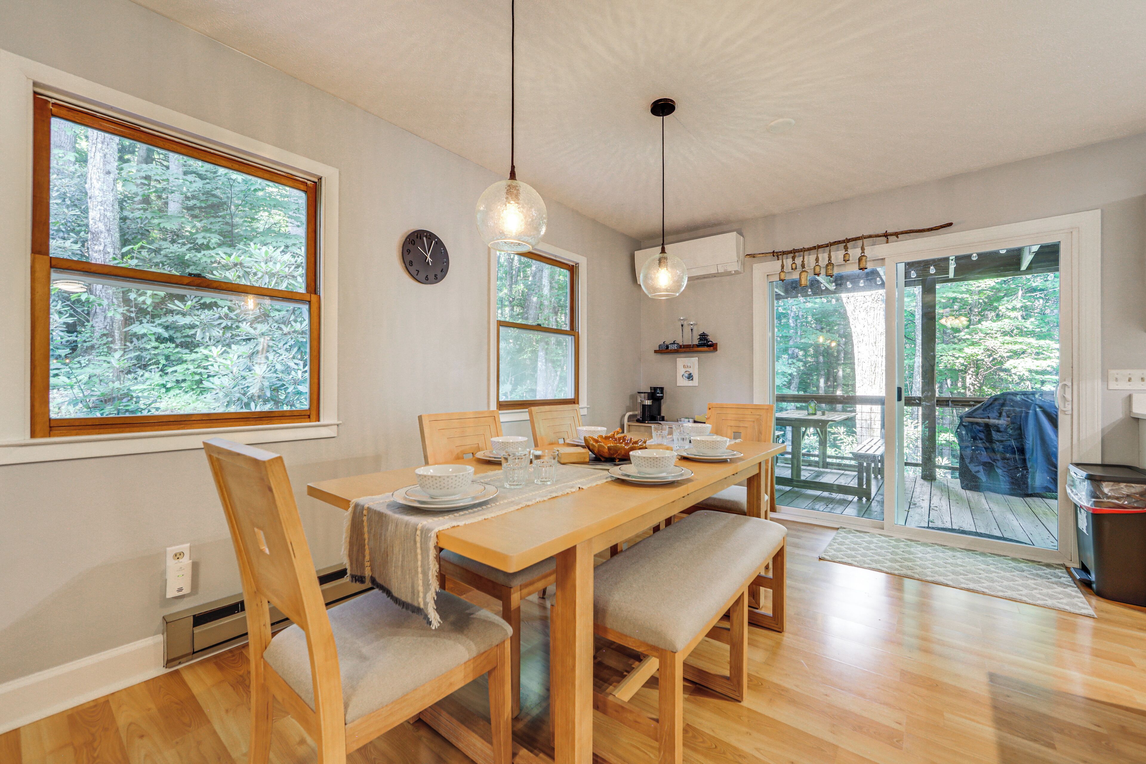 A dining area with a wooden table, chairs, and a view of a forest through large windows.