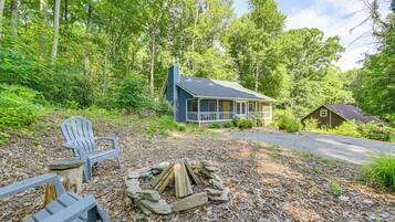 A cabin with a porch, surrounded by a forest. There are Adirondack chairs and a fire pit.