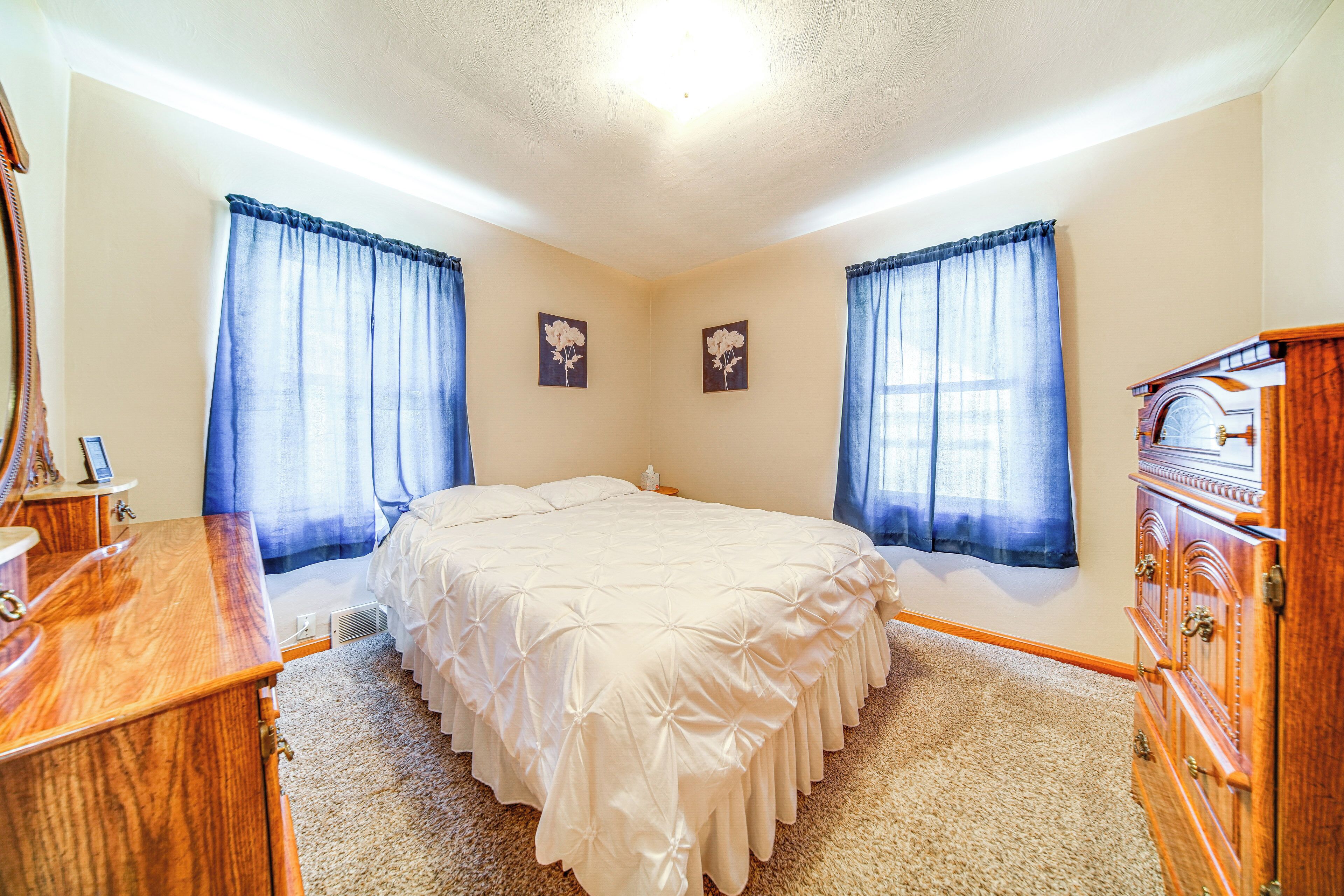 A bedroom with a bed, wooden dresser, and two windows with curtains.