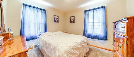 A bedroom with a bed, wooden dresser, and two windows with curtains.
