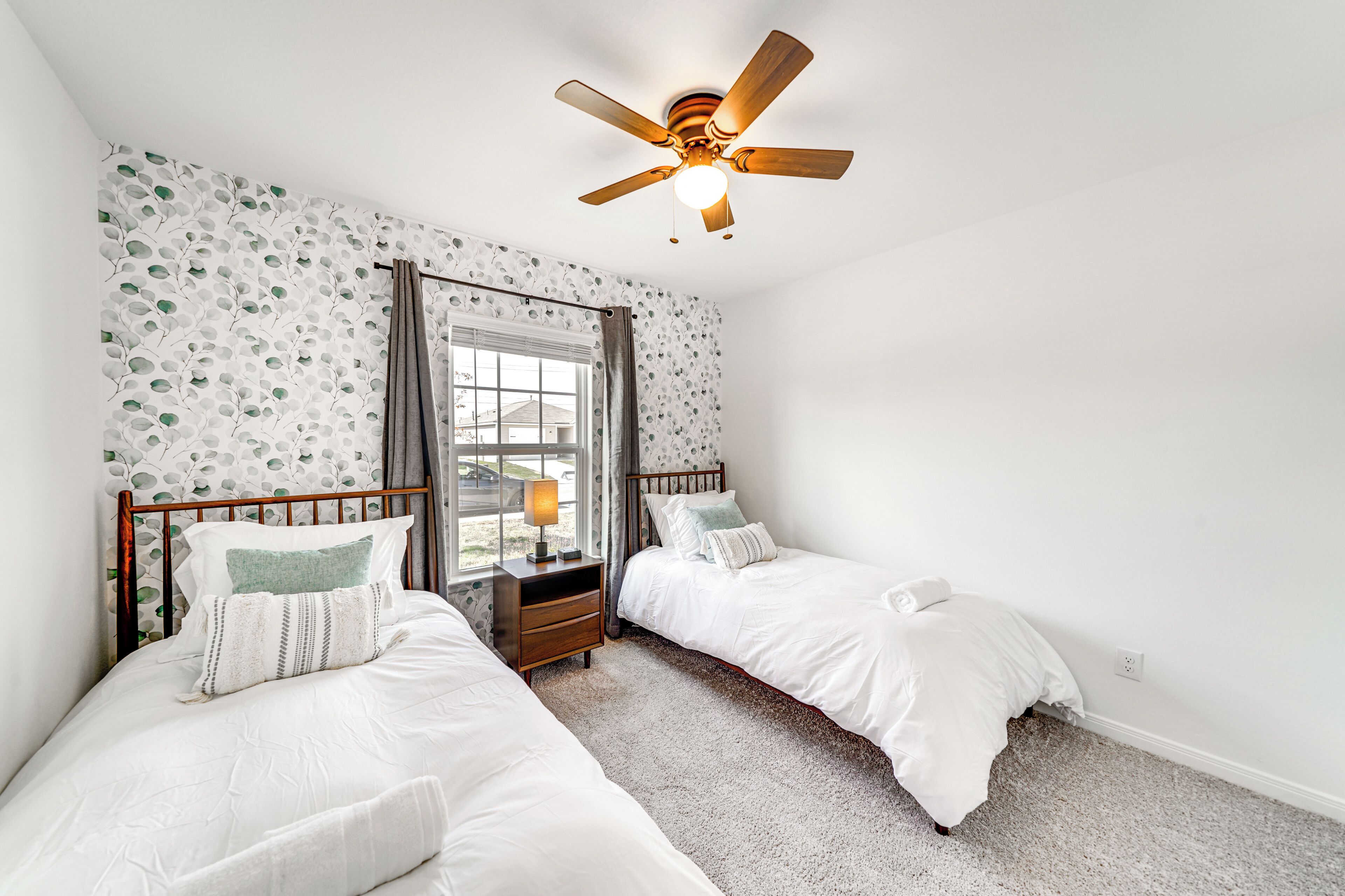 Un dormitorio con dos camas, un ventilador de techo, una ventana con cortinas y una mesita de noche con lámpara.