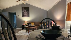 A dining area with a long table set for four, a wooden floor, a ceiling fan, and a view of the living room in the background.