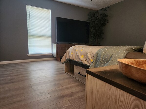 A bedroom with a bed, a TV, a wooden dresser, and a window with blinds.