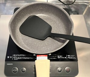 Una cocina con una sartén antiadherente sobre una placa de inducción, una espátula y un panel de control con texto en japonés.