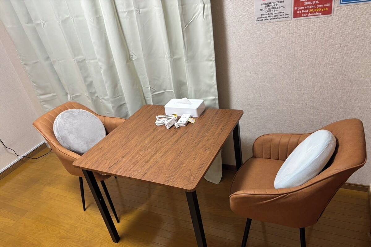 A small room with a wooden table and two brown chairs with cushions. There is a white box on the table and a power strip with cords.