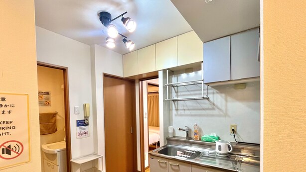Una cucina compatta con lavandino, piano di lavoro e pensili. Si nota un cartello appeso al muro e una porta che dà su un'altra stanza.