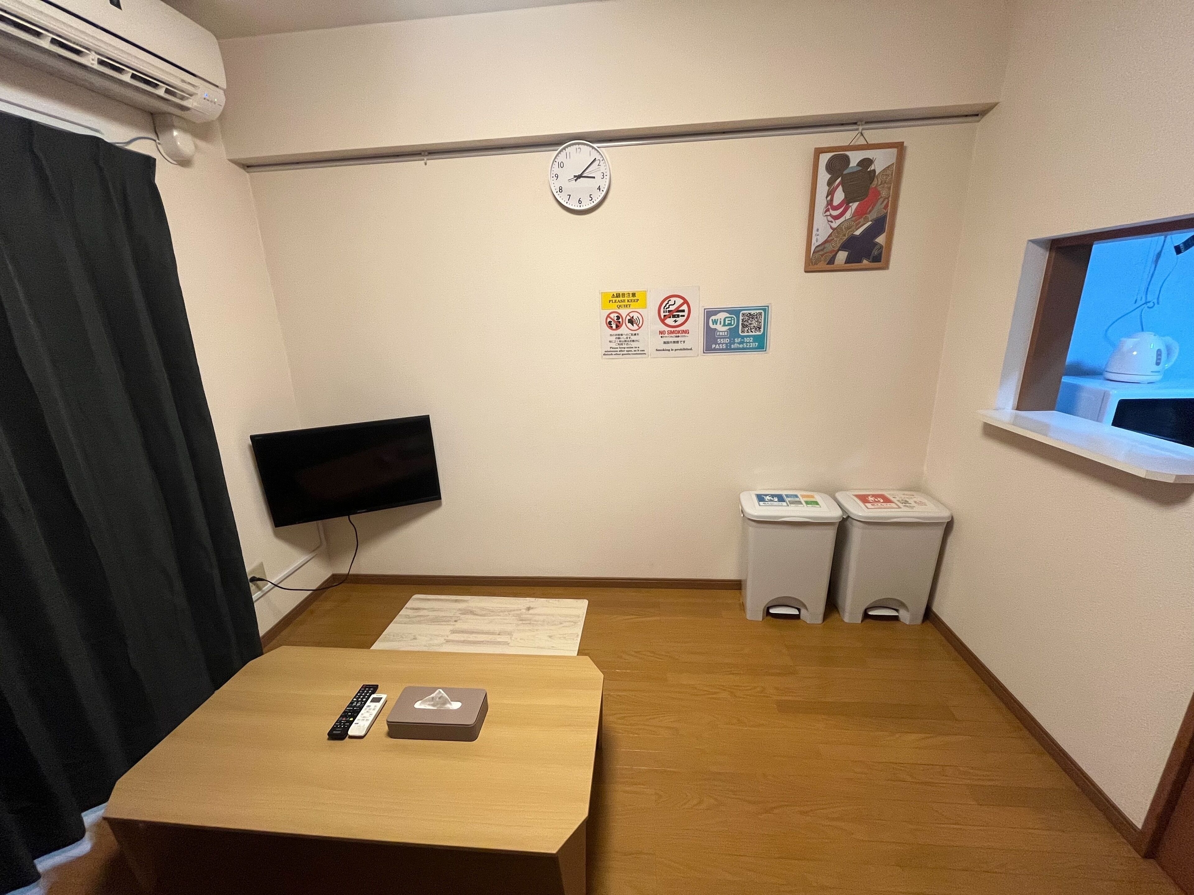 A hotel room with a wooden floor, a flat-screen TV mounted on the wall, a clock, a window with a view, and a small table with a remote control and a box.