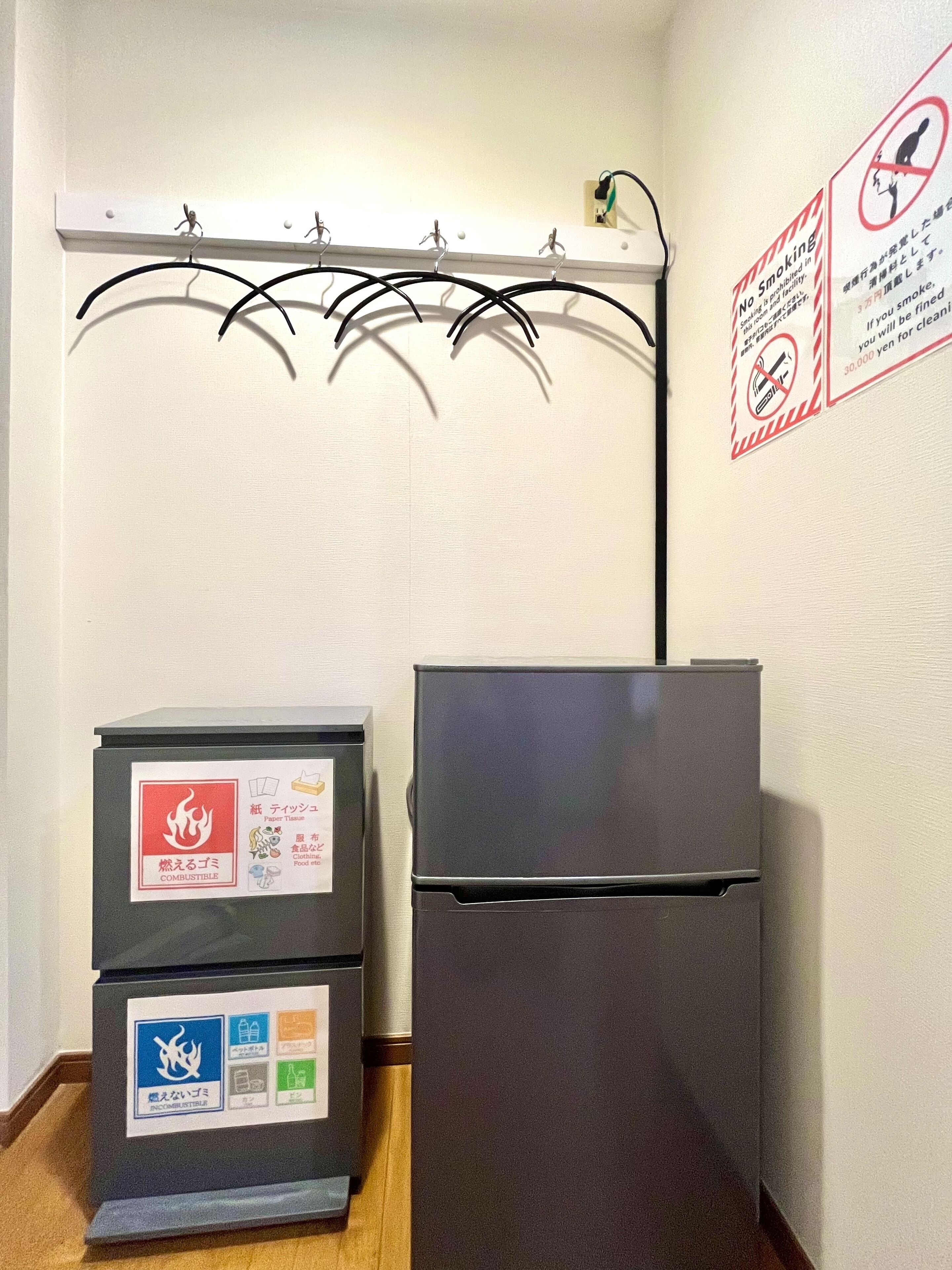 A small room with a black refrigerator, a trash bin, and a coat rack with no items on it. There are two signs on the wall, one prohibiting smoking and another warning about combustible materials.