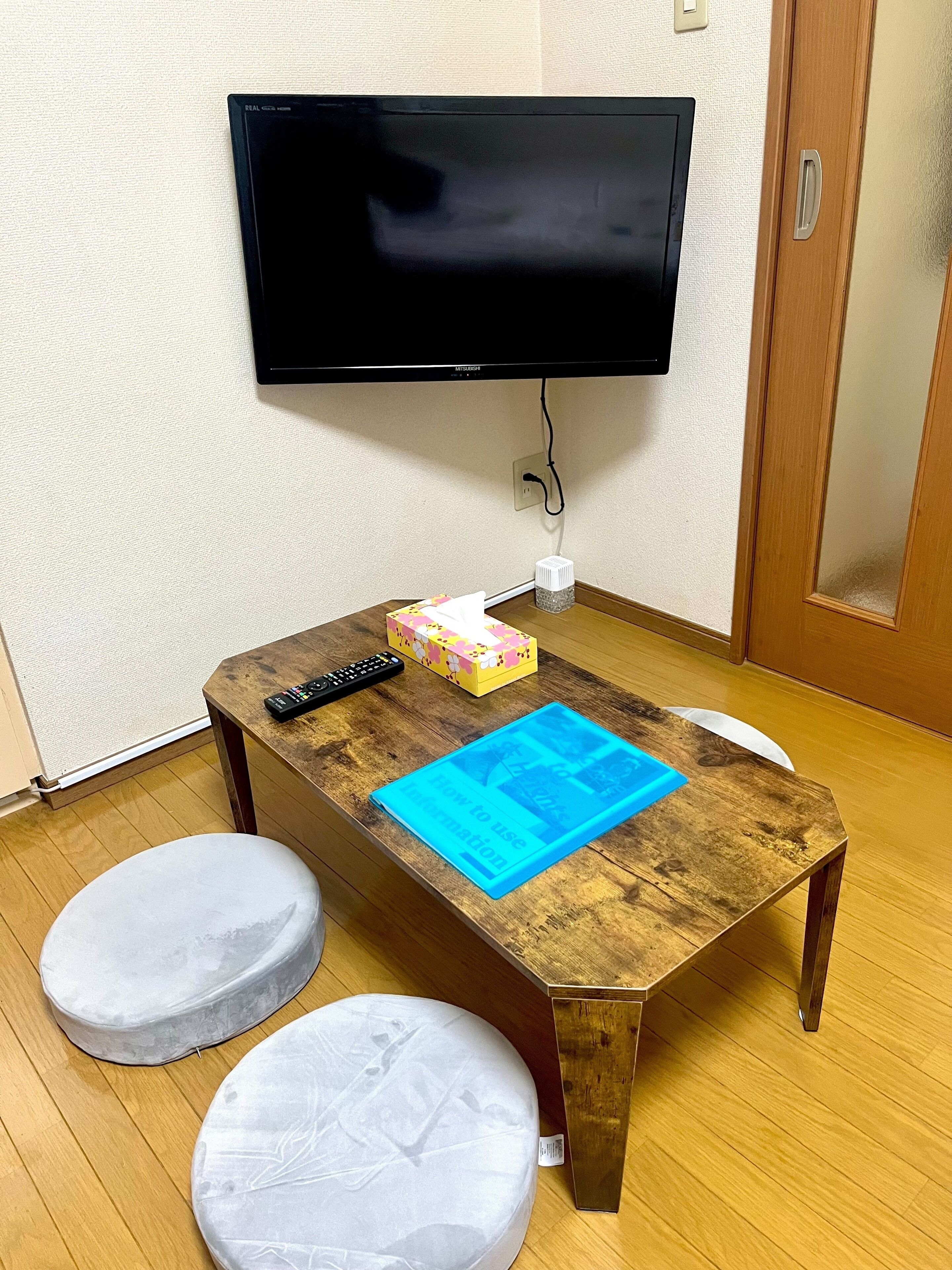 A small room with a wooden table, two cushioned stools, a television mounted on the wall, and a door.